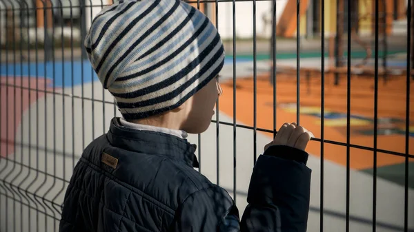 Molesto niño estresado sosteniendo y mirando a través de valla de metal alto en la escuela y el patio caro. Concepto de pobreza, inmigración, bullying y estrés infantil — Foto de Stock