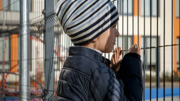 Menino perturbado e estressado olhando através da cerca de metal da escola depois foi intimidado. Conceito de pobreza, imigração, bullying e estresse infantil — Fotografia de Stock