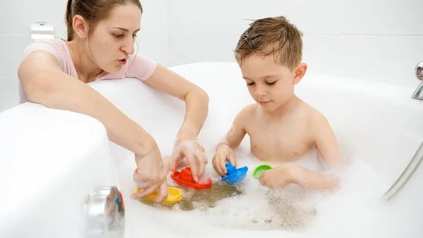 Lächelnde Mutter beim Spielen von Spielzeugbooten und Schiffen mit ihrem Sohn beim Waschen in der Badewanne. Konzept Familienzeit, Entwicklung der Kinder und Spaß zu Hause. — Stockfoto