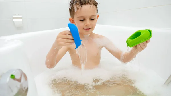 Веселий усміхнений хлопчик грає з іграшковими човнами і бризкає воду у ванній кімнаті. Концепція сімейного часу, розвиток дітей та веселощі вдома — стокове фото
