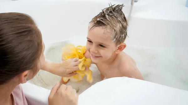 Молода любляча мати миє свого усміхненого сина з милом і губкою у ванній — стокове фото