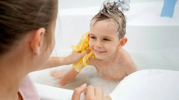 Joyeux garçon souriant qui regarde sa mère pendant qu'elle le lave avec du savon et une éponge dans la salle de bain. Concept d'hygine, développement des enfants et plaisir à la maison — Photo