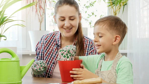 Молодая мать помогает сыну положить садовые перчатки, прежде чем сажать цветы в горшки в доме. Концепция садоводства, посадки на дому и совместной работы семьи. — стоковое фото