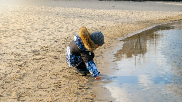 Rüzgarlı kış gününde, parkta soğuk suya dokunan mutlu çocuk. — Stok fotoğraf