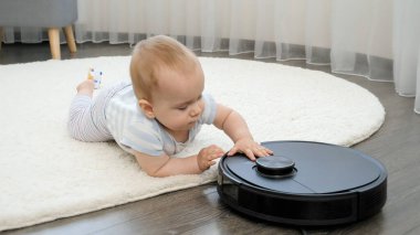 Küçük bebek yerde yatıp robot elektrik süpürgesiyle oynuyor.