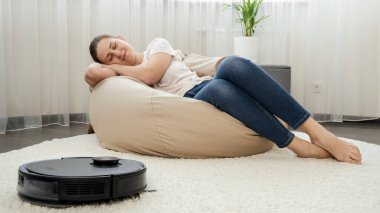 Oturma odasında çalışan robot elektrik süpürgesinin yanında uyuyan genç kadın. Modern hayatta hijyen, ev aletleri ve robotlar kavramı.