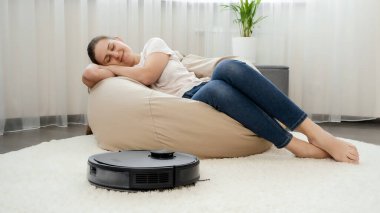 Robot elektrikli süpürge oturma odasındaki tozları temizlerken güzel, gülümseyen bir kadın koltukta uyuyor.
