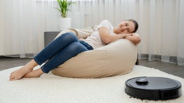 Mutlu, gülümseyen kadın sandalyede uzanıp rahatlıyor ve çalışan robot süpürgeye bakıyor.