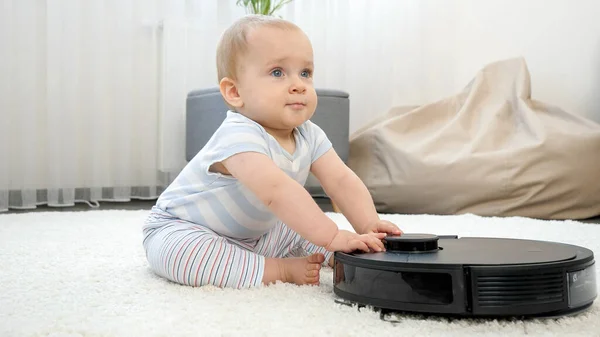 Glad leende pojke sitter på mattan och leker med robot dammsugare. Begreppet hygien, hushållsapparater och robotar i det moderna livet. — Stockfoto