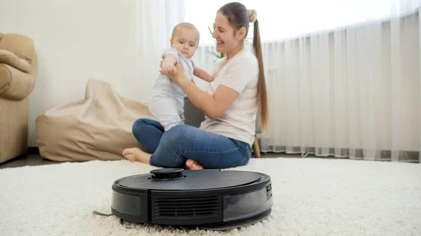 Robot aspirateur aider la femme à faire des tâches pendant qu'elle passe du temps avec bébé fils. Concept d'hygiène, gadgets ménagers et robots à la vie moderne. — Photo