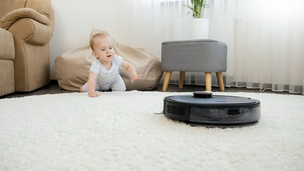 Robotdammsugare rör sig mot en liten pojke som kryper på golvet hemma. Begreppet hygien, hushållsapparater och robotar i det moderna livet. — Stockfoto