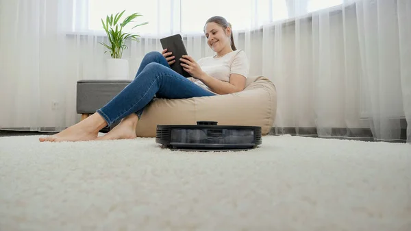 Gelukkig glimlachende vrouw ontspannen in stoel en surfen op internet op tablet computer, terwijl robot stofzuiger doet huishoudelijk werk en opruimen — Stockfoto