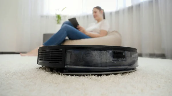 CLoseup de robot aspirateur nettoyage maison tandis que la jeune femme travaillant dans le salon. Concept d'hygiène, gadgets ménagers et robots à la vie moderne. — Photo