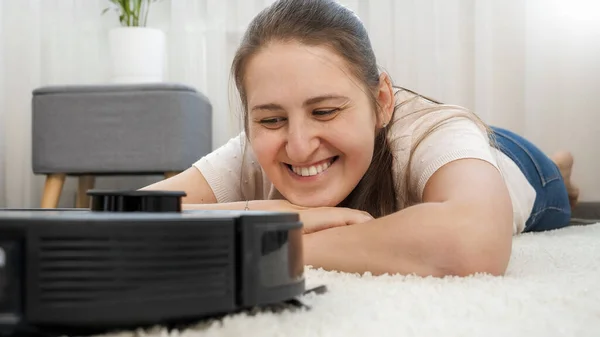 거실 카펫에 누워 행복 한 미소짓는 여성의 모습 과 일하는 로봇 청소기를 보고 있는 모습 — 스톡 사진