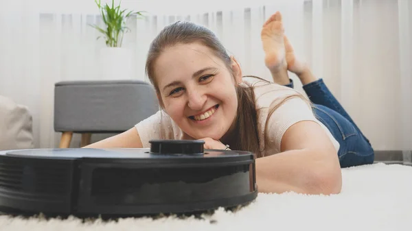Σκυμμένο πορτρέτο της χαρούμενης χαμογελαστής γυναίκας ξαπλωμένη στο χαλί δίπλα στο νέο ρομπότ καθαρίστρια που κάνει δουλειές του σπιτιού. — Φωτογραφία Αρχείου