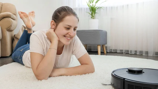 Gelukkige lachende vrouw met werkende robot stofzuiger op overstroming in de woonkamer. Concept van hygiëne, huishoudelijke apparaten en robots in het moderne leven. — Stockfoto