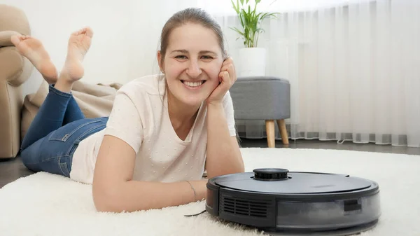 Portret van een lachende vrouw die naar haar nieuwe robot stofzuiger kijkt die in de woonkamer werkt. Concept van hygiëne, huishoudelijke apparaten en robots in het moderne leven. — Stockfoto