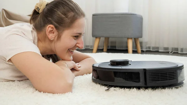 Detailní záběr vysavače robotů míří k usměvavé ženě ležící na koberci v obývacím pokoji. Koncepce hygieny, domácích pomůcek a robotů v moderním životě. — Stock fotografie