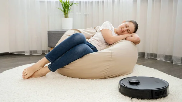 Jonge huisvrouw ontspannen in de woonkamer, terwijl robot stofzuiger doet huishoudelijk werk. Concept van hygiëne, huishoudelijke apparaten en robots in het moderne leven. — Stockfoto