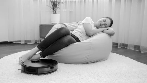 Imagen en blanco y negro de una mujer joven relajada en una silla mientras que la aspiradora robot hace las tareas domésticas y la limpieza — Foto de Stock