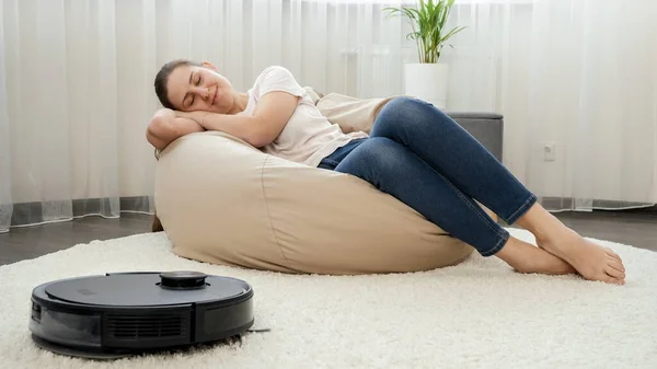 Young woman sleeping in living room next to working robot vacuum cleaner. Concept of hygiene, household gadgets and robots at modern life. — Stock Photo, Image