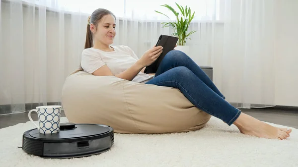 Jonge freelance vrouw die thuis werkt en koffie drinkt van een robot stofzuiger. Concept van hygiëne, huishoudelijke apparaten en robots in het moderne leven. — Stockfoto