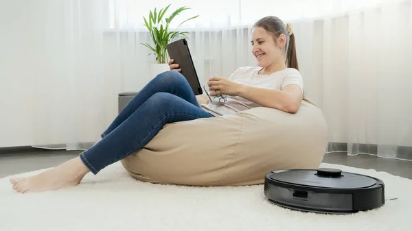 Glad leende kvinna avkopplande i stol med kopp te och surfplatta dator medan robot dammsugare gör hushållsarbete och rengöring mattan — Stockfoto
