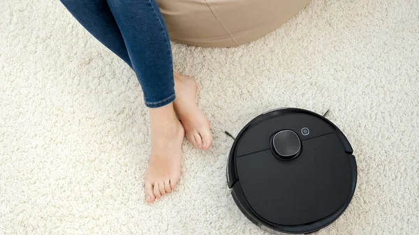 Staubsaugerroboter reinigen Teppich um barfüßige Frau, die im Stuhl sitzt. Hygienekonzept, Haushaltsgeräte und Roboter im modernen Leben. — Stockfoto