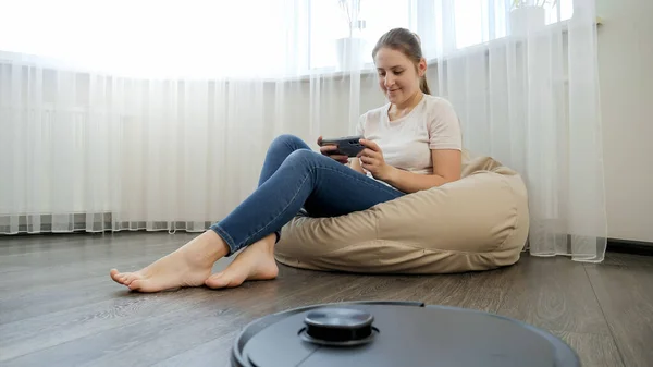 Robot dammsugare gör hushållsarbete medan ung kvinna koppla av och vila med smartphone i Beanbag stol. Begreppet hygien, hushållsapparater och robotar i det moderna livet. — Stockfoto