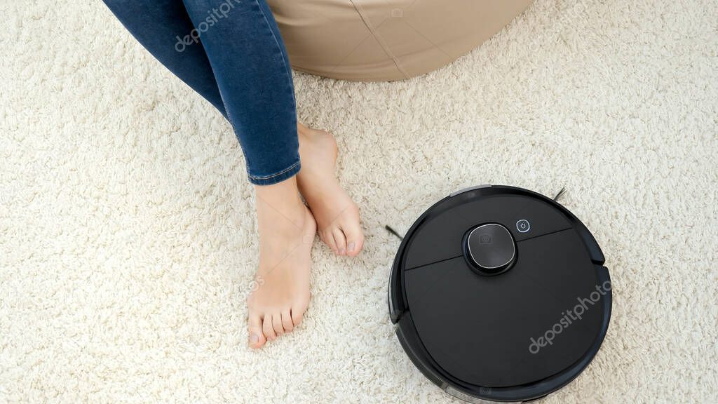 Robot vacuum cleaner cleaning carpet around barefoot woman sitting in chair. Concept of hygiene, household gadgets and robots at modern life.