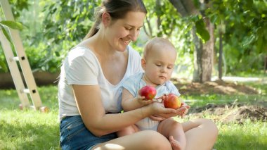 Annesi meyve bahçesinde kırmızı elma tutan tatlı, gülümseyen bir çocuk. Çocuk gelişimi, ebeveynlik ve aile kavramı açık havada birlikte dinleniyor..