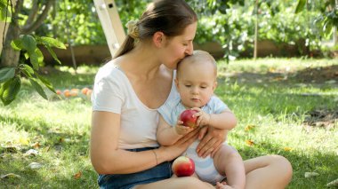 Genç, şefkatli bir anne, küçük oğlunu öpüyor. Evcil bahçede kırmızı olgun bir elma tutuyor.