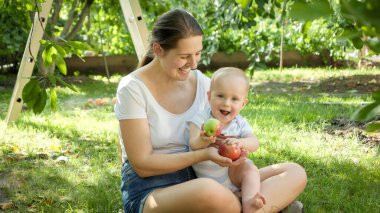 Genç bir anne, evinin arka bahçesinde elinde olgun kırmızı elmalarla küçük oğluna sarılıp gülümsüyor. Çocuk gelişimi, ebeveynlik ve sağlıklı organik gıda yetiştirme kavramı.