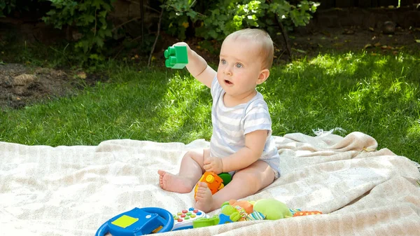 Szczęśliwy uśmiechnięty chłopiec siedzący na kocu w parku i bawiący się plastikowymi zabawkami. Koncepcja wczesnego rozwoju dziecka, edukacji i relaksu na świeżym powietrzu. — Zdjęcie stockowe