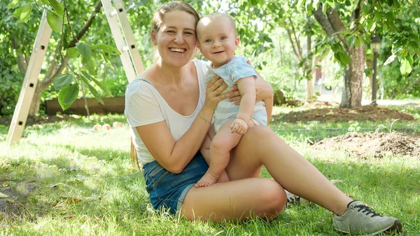 Portret van een vrolijk lachend jongetje, zittend met moeder op gras onder appelbomen en kijkend in de camera — Stockfoto