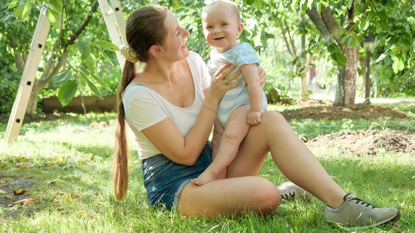 Cheerful baby boy sitting on grass at apple orchard with young mother — Stock Photo, Image