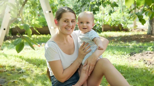 Retrato da mãe sorridente feliz segurando e beijando seu filho bebê enquanto relaxa sob a macieira no jardim. Conceito de desenvolvimento precoce da criança, educação e relaxamento ao ar livre. — Fotografia de Stock