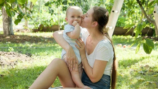Gelukkig jong moeder knuffelen en zoenen haar baby zoon onder bomen in tuin — Stockfoto