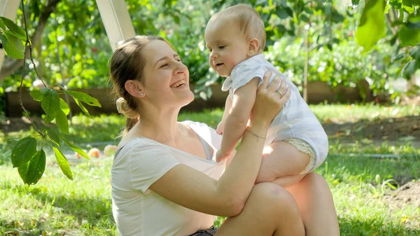 Portret van een lachende liefhebbende moeder die haar zoontje knuffelt terwijl ze zich ontspant op gras in de tuin. Concept van de ontwikkeling van het kind, familie die samen tijd doorbrengt en ouderschap. — Stockfoto