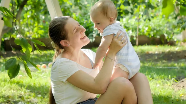 Retrato de mãe feliz segurando e olhando para seu filho bebê sob árvores no jardim — Fotografia de Stock