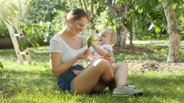 Mladá milující matka dává strom list na své malé dítě syna, zatímco sedí na trávě na zahradě — Stock fotografie