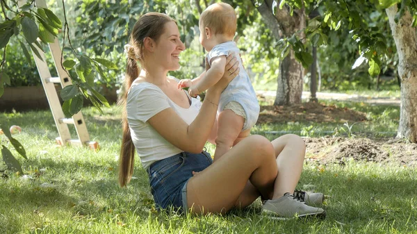 Madre sonriente dando hojas de árbol a su pequeño hijo mientras está sentada en la hierba bajo el manzano en el huerto. Concepto de desarrollo infantil, pasar tiempo en familia y crianza de los hijos. — Foto de Stock