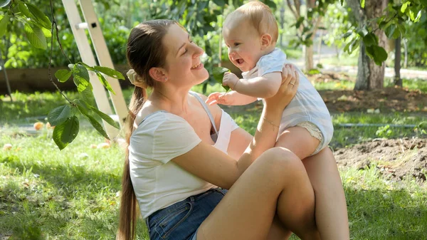 Retrato de una mujer sonriente sentada en la hierba y sosteniendo a su pequeño hijo en el soleado día de verano — Foto de Stock