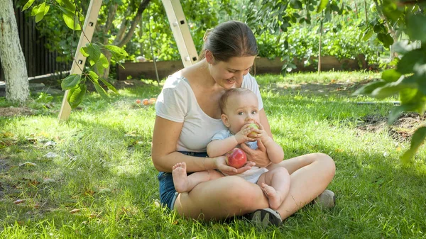 Lächelnde junge Frau sitzt mit ihrem kleinen Sohn im Garten und schaut ihn beim Äpfelessen an — Stockfoto
