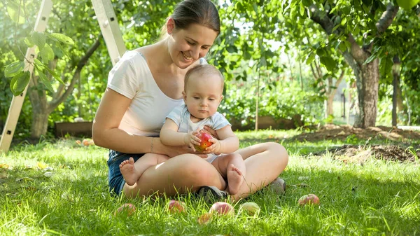 Felice madre sorridente con il piccolo bambino seduto a frutteto e raccogliendo mele mature. Concetto di sviluppo del bambino, genitorialità e famiglia rilassarsi insieme all'aperto. — Foto Stock