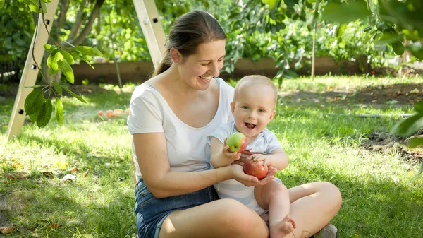 Joven madre abrazando y sonriendo a su hijo bebé sosteniendo manzanas rojas maduras en el jardín del patio trasero de la casa. Concepto de desarrollo infantil, crianza y cultivo de alimentos orgánicos saludables. — Foto de Stock