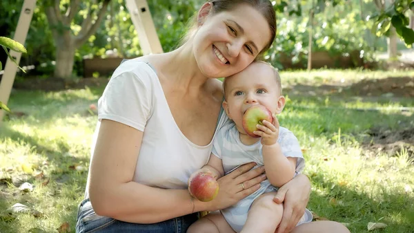Porträt einer lächelnden Mutter, die mit ihrem kleinen Sohn im Garten Äpfel auf Gras isst — Stockfoto