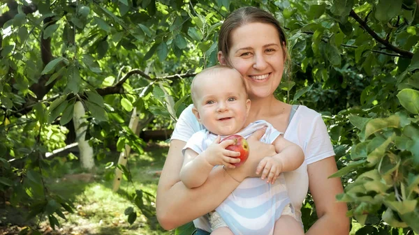 Portret van een lachende baby met moeder die rijpe appels vasthoudt en poseert in de tuin. Concept van kinderontwikkeling, ouderschap en gezonde biologische voeding. — Stockfoto