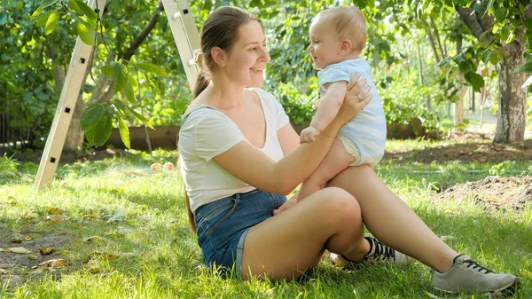 Glücklich lächelnde Mutter hebt ihren kleinen Sohn auf und küsst ihn unter Apfelbäumen im Obstgarten. Konzept der frühkindlichen Entwicklung, Bildung und Entspannung im Freien. Stockbild