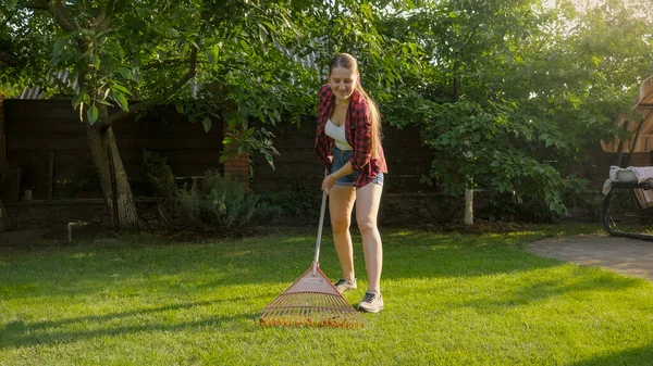 Jonge lachende vrouw die in de achtertuin werkt en gevallen bladeren verzamelt met hark. Begrip huishoudelijk werk, tuinieren en stadsleven — Stockfoto